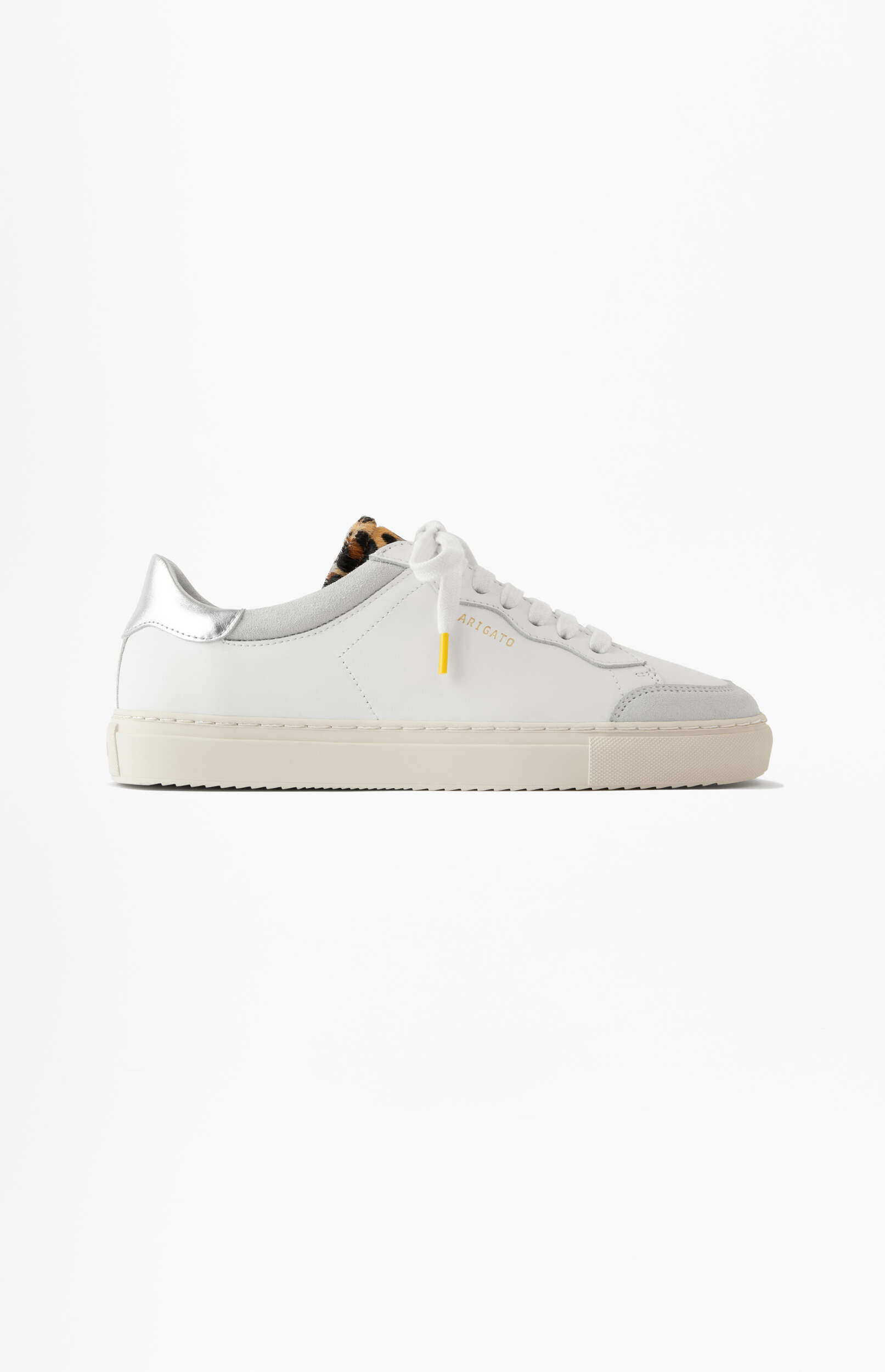 A white leather sneaker with a silver heel tab, a subtle leopard print lining visible at the top, and 'ARIGATO' embossed in gold on the side. The laces are white with a yellow tip.