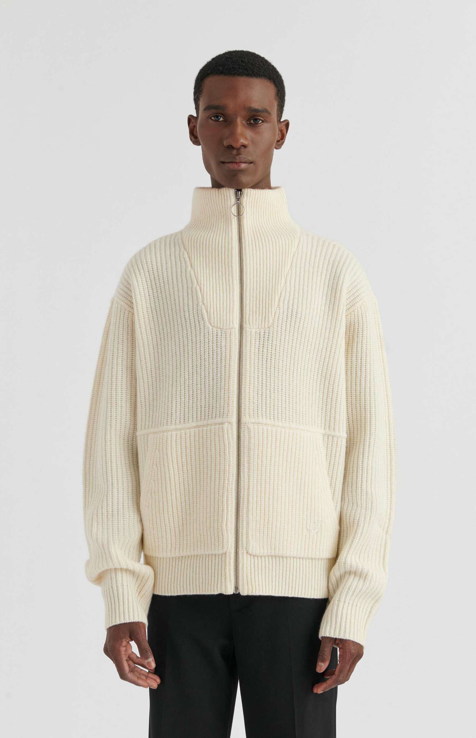 A man with dark skin and short dark hair is shown from the waist up, wearing a cream-colored, ribbed knit zip-up sweater over black pants, against a plain white background. The sweater has a high neck, a silver zipper, and two prominent square patch pockets.