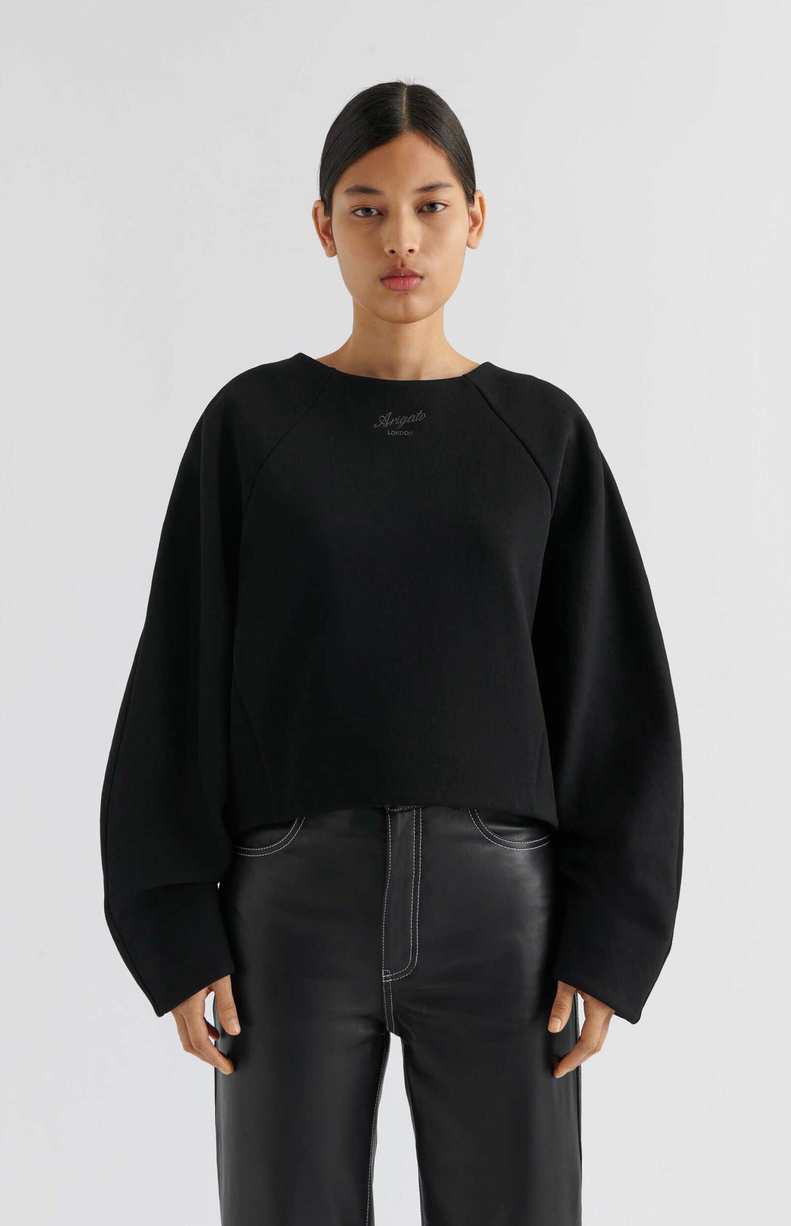 A model standing against a white background, wearing a black sweatshirt with 'Arigato London' embroidered on the chest and black leather trousers with white contrasting stitching.