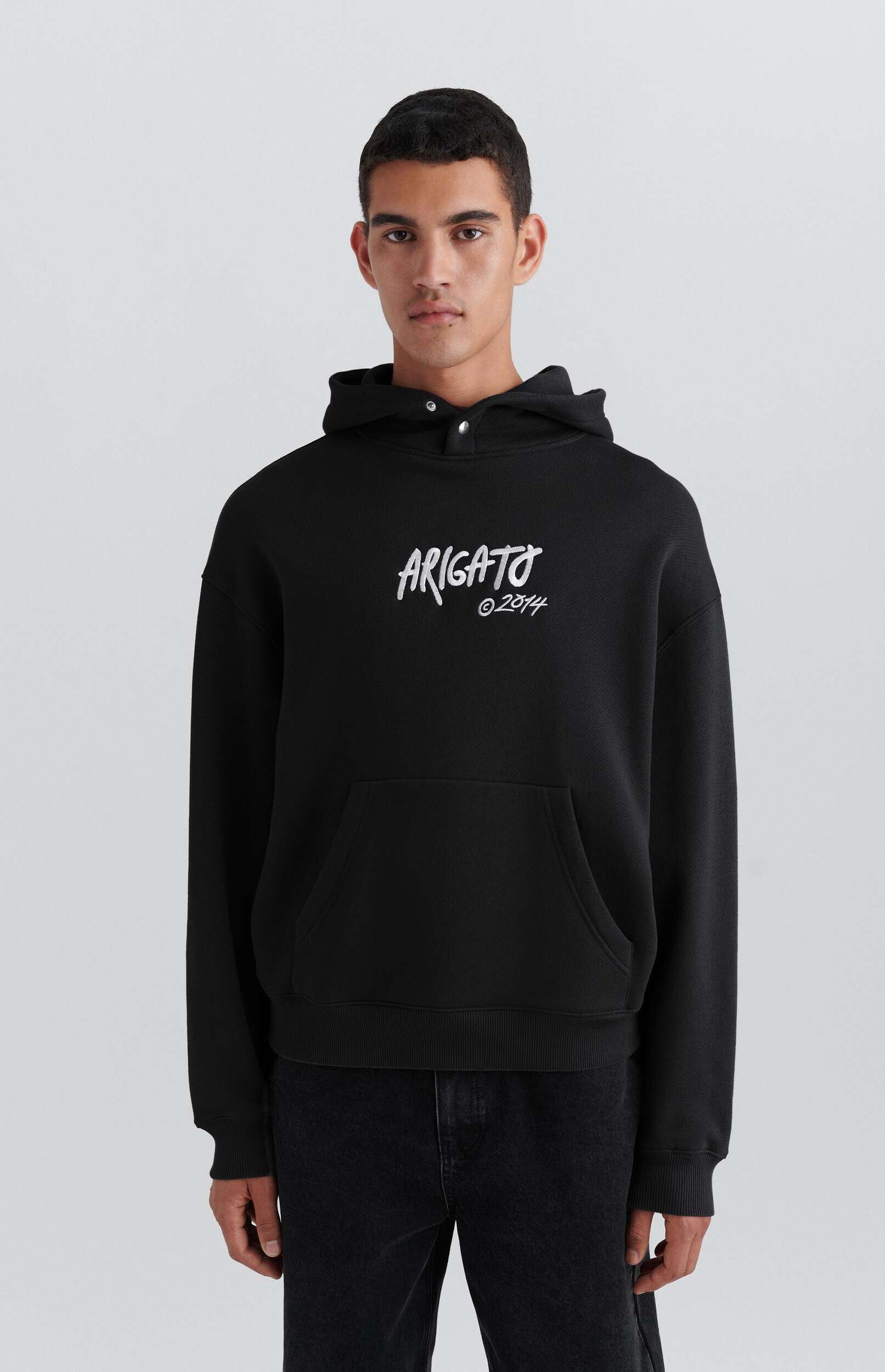 A man stands facing forward, modeling a black hoodie with 'ARIGATO' in white script and '© 2014' below it, paired with black jeans, against a plain white background.