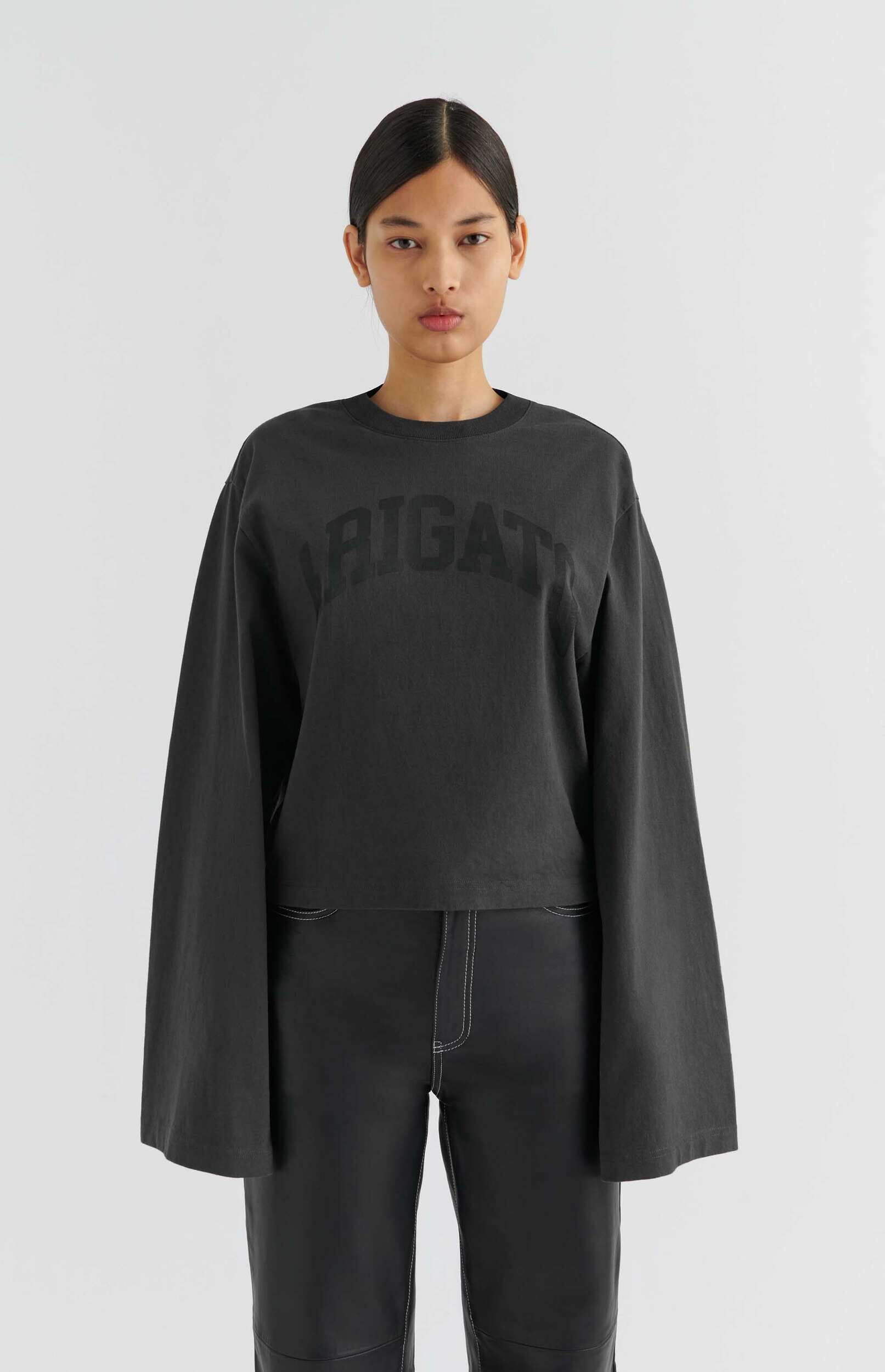 A full-body shot of a person with dark hair pulled back, wearing a dark gray oversized long-sleeve sweatshirt with wide, flared sleeves and a subtle 'RIGAT' logo on the chest, paired with dark pants featuring prominent white contrast stitching. The person stands against a plain white background, looking directly at the camera with a neutral expression.