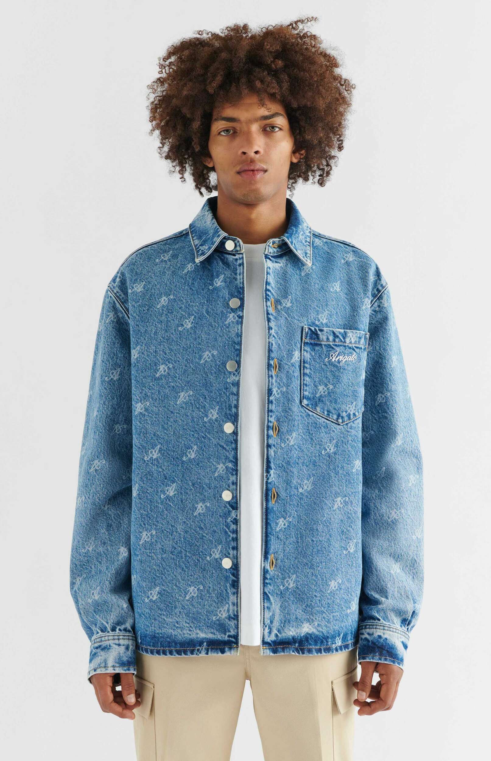 A man with curly hair wearing a patterned blue denim jacket over a white t-shirt and light beige pants, posing against a white background.