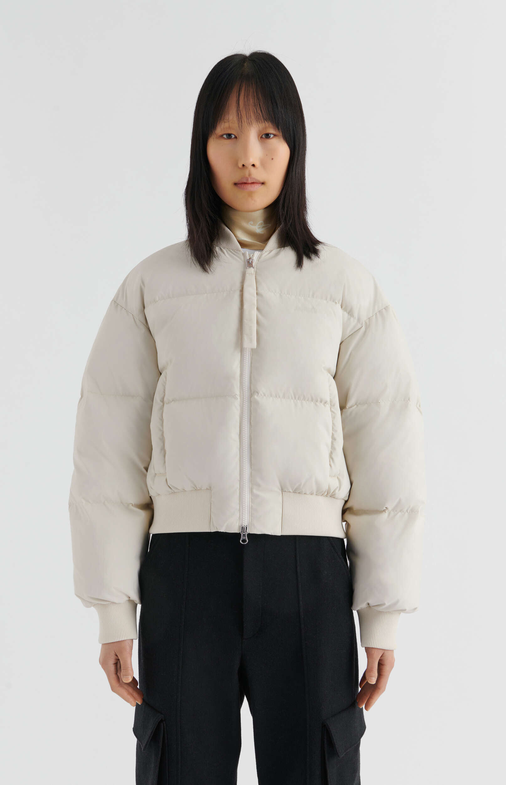 A young woman with straight black hair and bangs stands facing forward against a plain white background, wearing a light beige or off-white cropped puffer jacket with ribbed cuffs and hem, zipped up over a lighter turtleneck, and black cargo-style pants. Her expression is neutral.