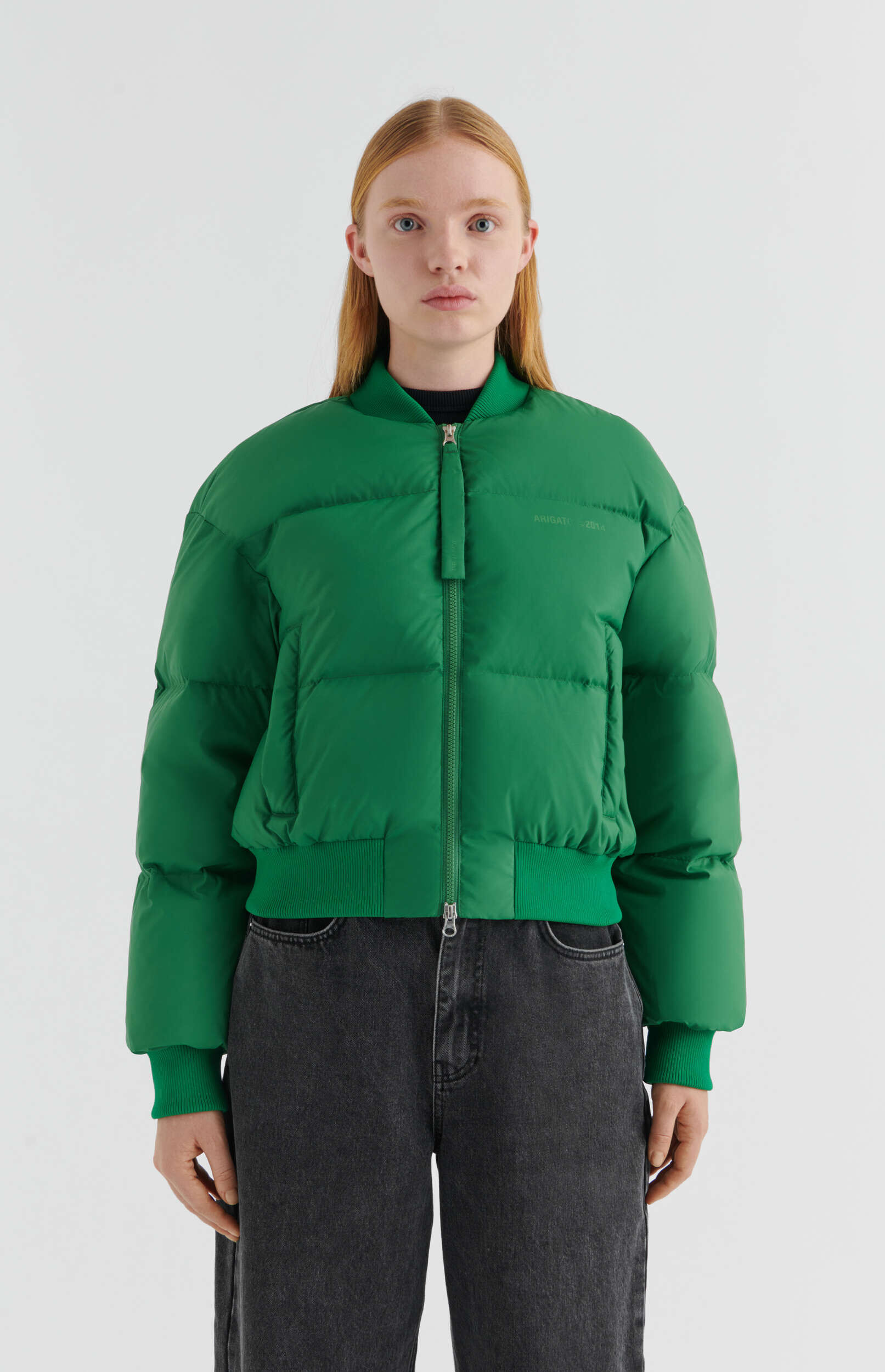 A young woman with light eyes and long reddish-blonde hair stands front-facing against a plain white background, wearing a vibrant green puffer bomber jacket with a front zipper and ribbed collar, cuffs, and hem, paired with dark grey relaxed-fit jeans.