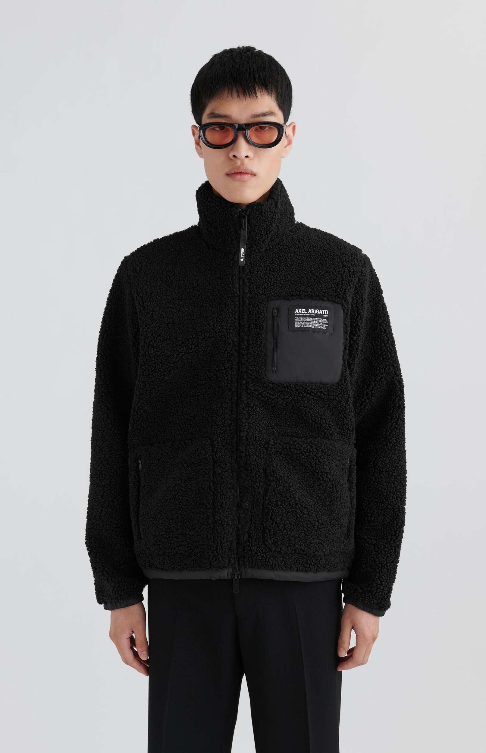 A person with short dark hair and orange-tinted sunglasses wears a black Axel Arigato high-collar sherpa jacket with a zippered chest pocket and black pants, posing against a light background.