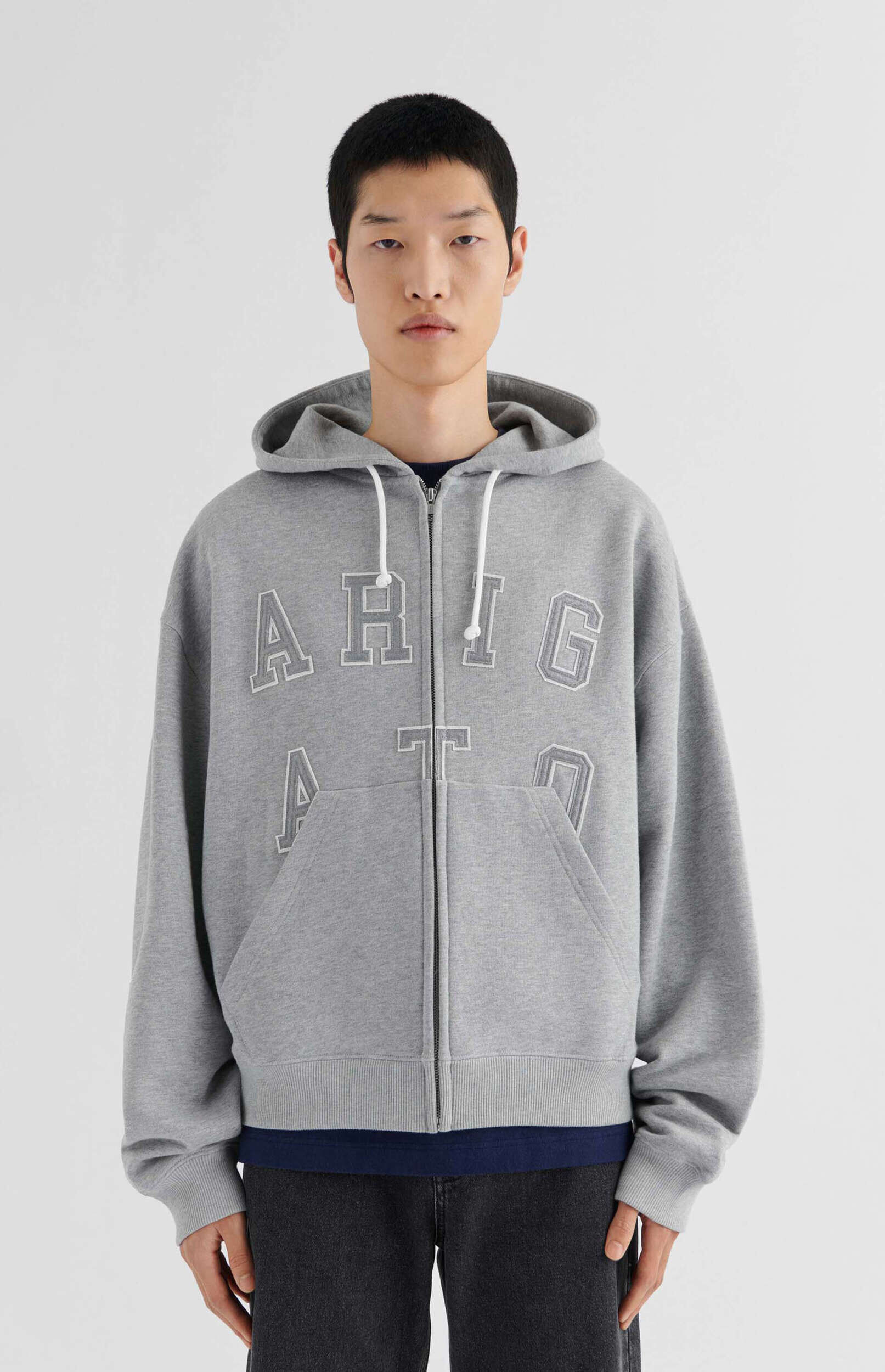 A young man with short black hair and an earnest expression stands front-facing, modeling a light grey zip-up hoodie with 'ARIGATO' in large, outlined white letters across the chest. He wears a dark blue crew-neck t-shirt underneath and dark grey jeans, with his hands out of view. The background is a plain white studio setting.