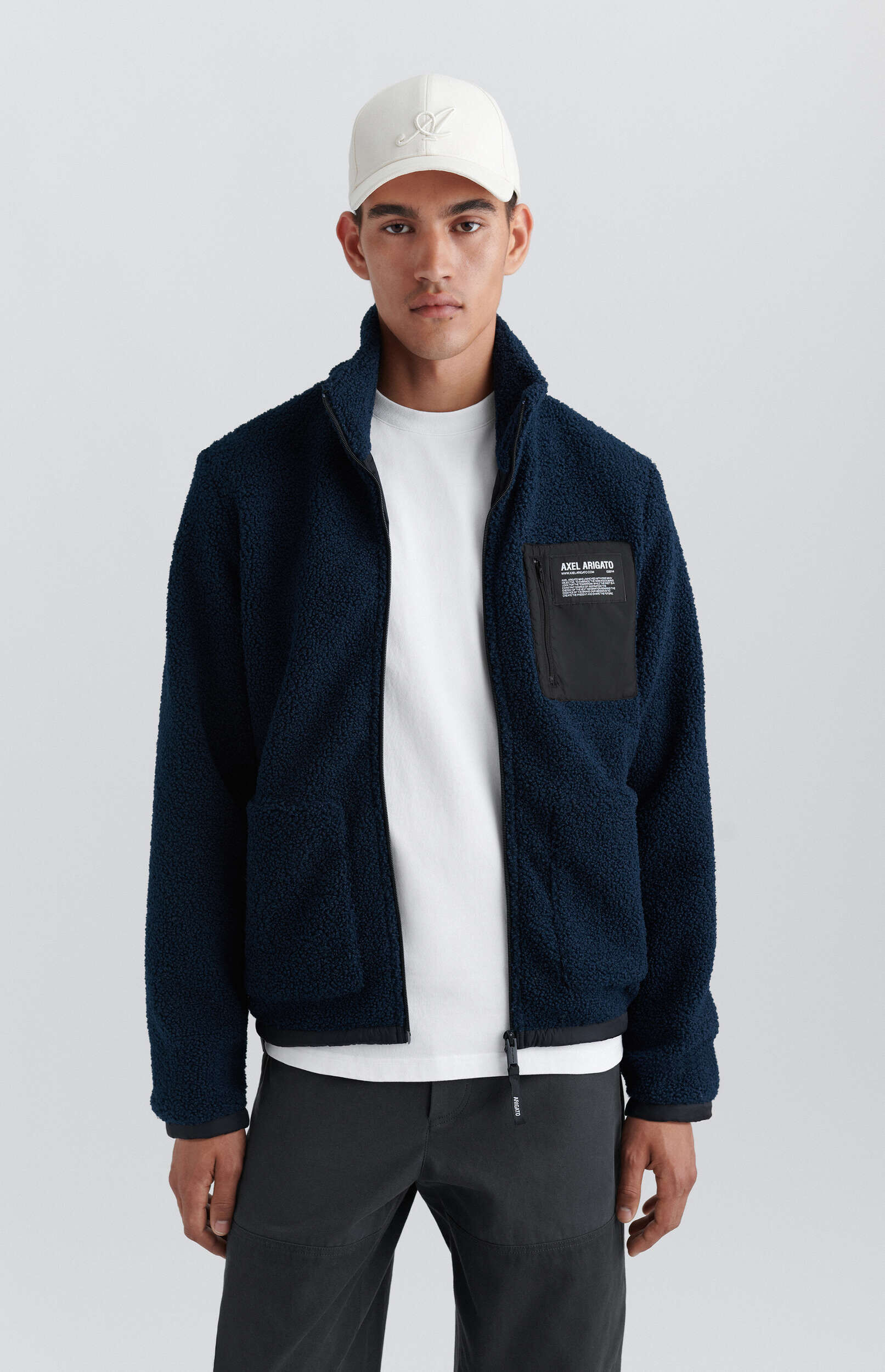 A young man wearing a white baseball cap, a navy blue sherpa fleece jacket with a black zippered chest pocket featuring the 'AXEL ARIGATO' logo, a white t-shirt, and dark gray pants, poses against a light gray background.