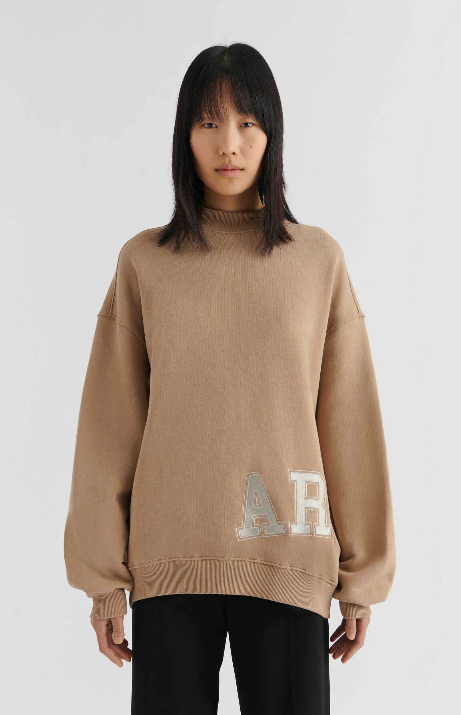 A young person with straight black hair and bangs wearing a loose-fitting, high-neck tan sweatshirt with large, silver, embossed letters 'AR' on the front, paired with black pants, standing against a plain white background.