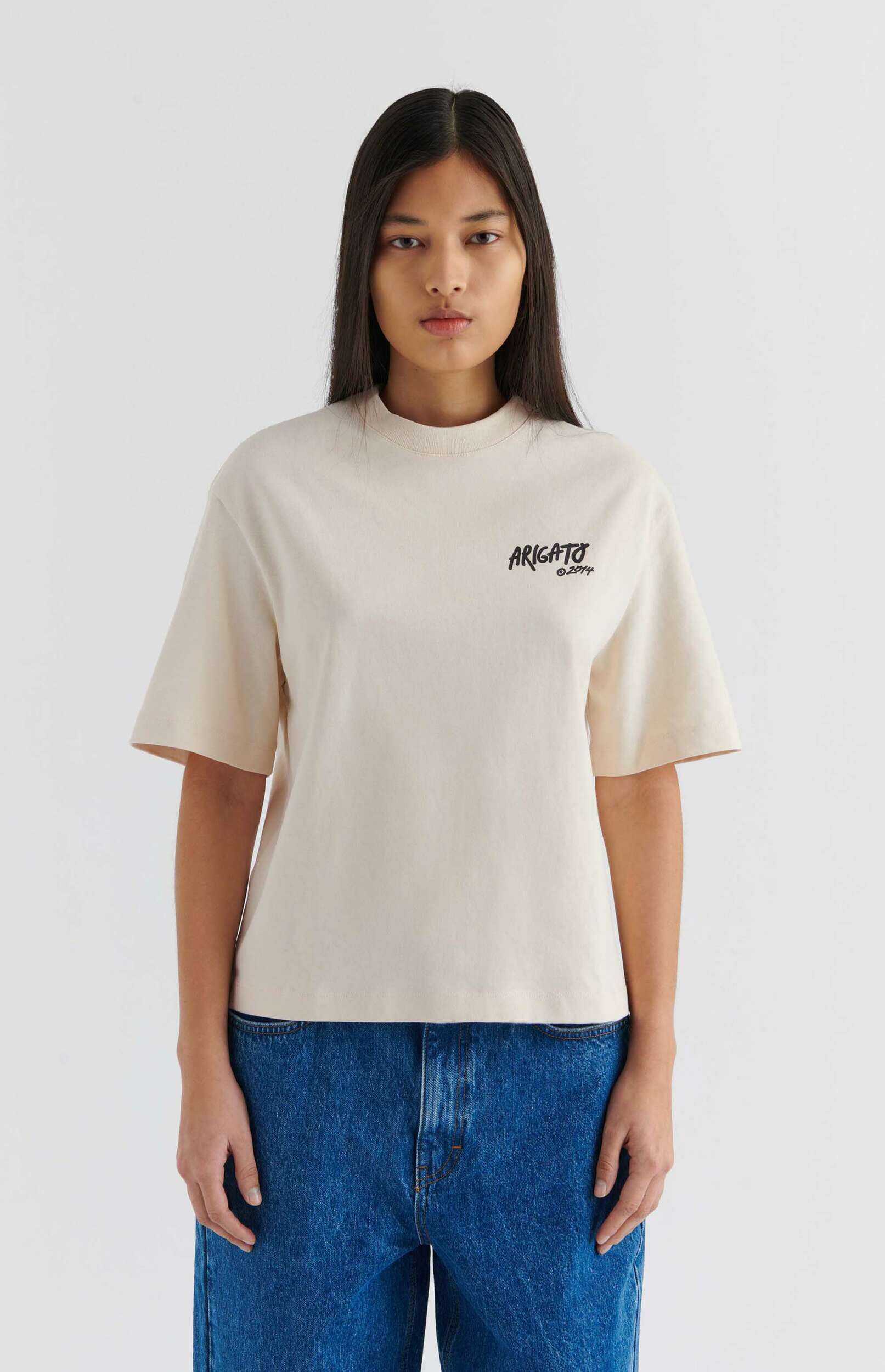 A young woman with long dark hair stands against a white background, wearing a cream t-shirt with 'ARIGATO 
A young woman with long dark hair stands against a white background, wearing a cream t-shirt with "ARIGATO 
©2014" printed on the chest, and blue denim jeans.
