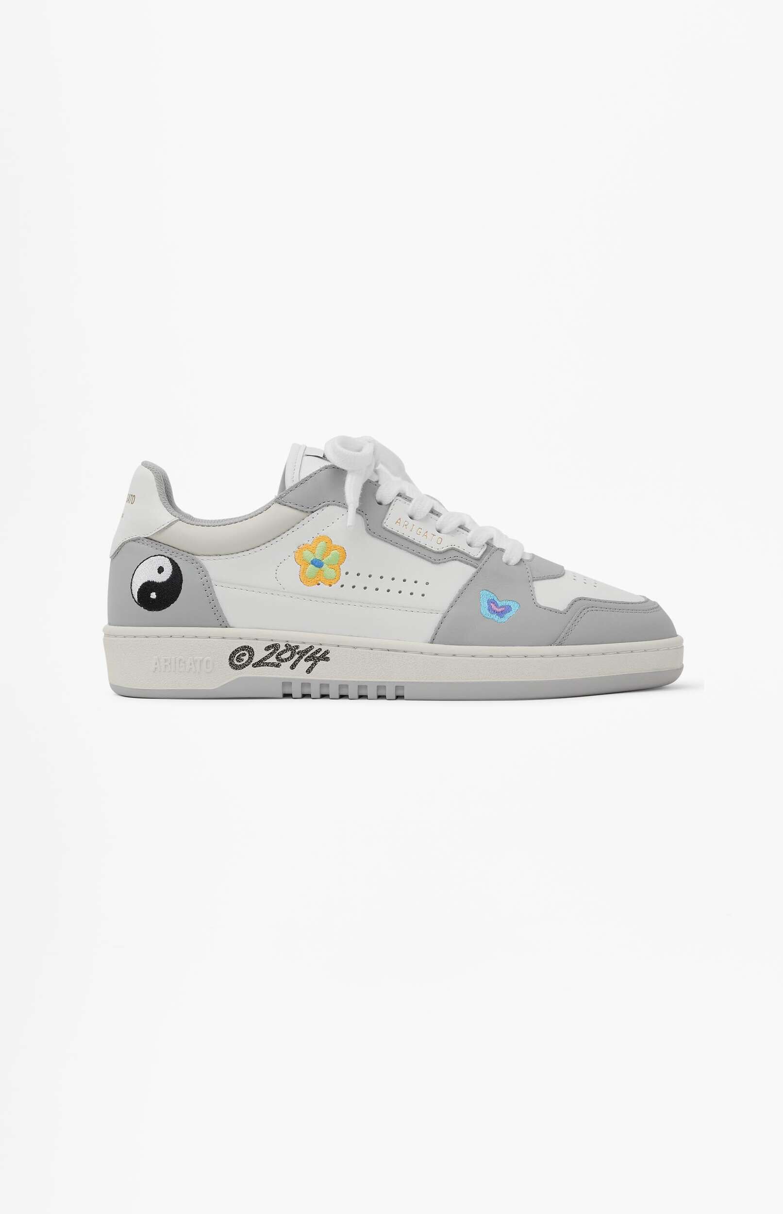 A white and grey low-top sneaker featuring a black and white yin-yang symbol on the heel, a yellow and orange embroidered flower, and a light blue embroidered butterfly on the sides. The brand 'ARIGATO' and '©2014' are visible on the sole.