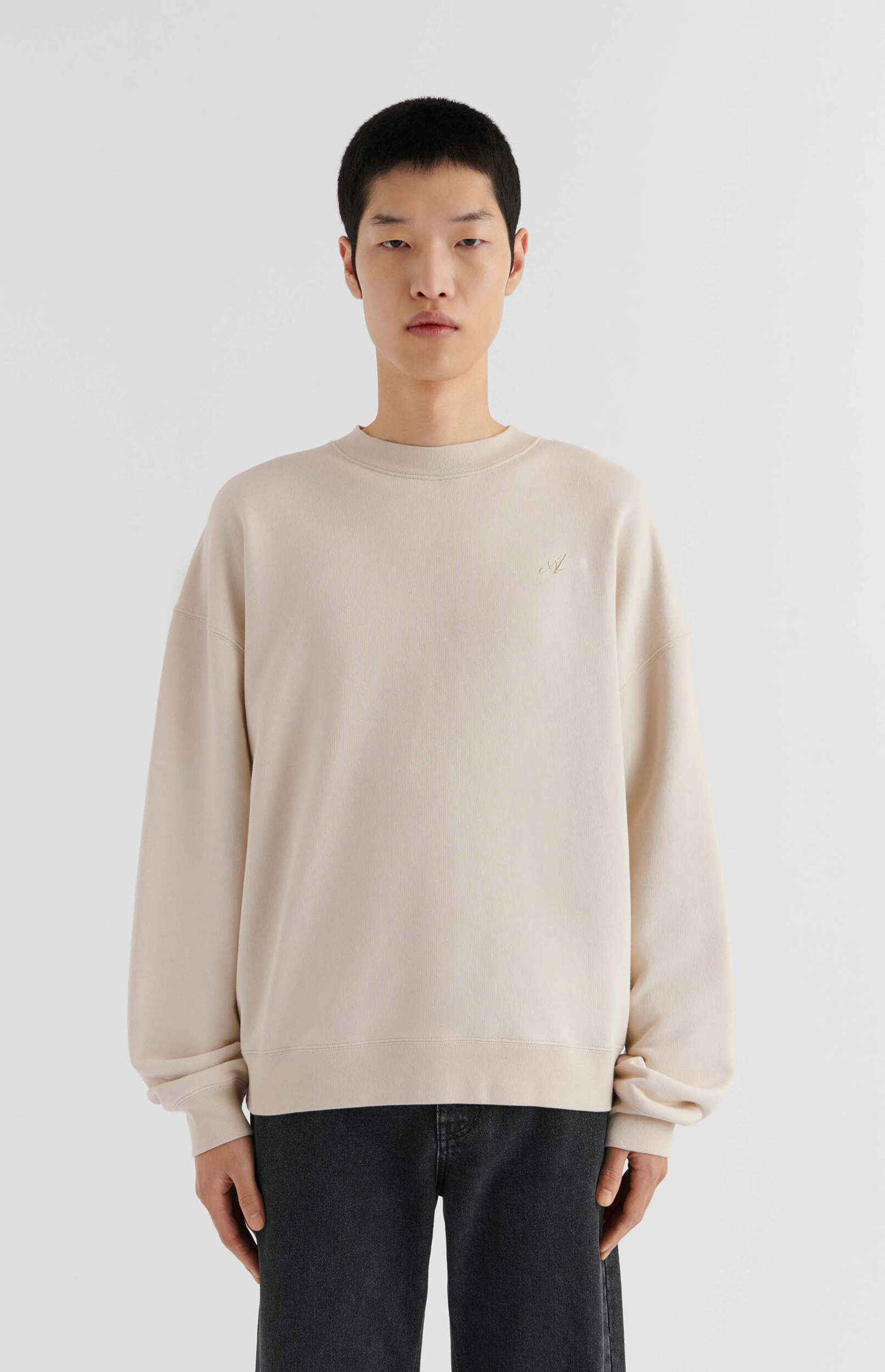 A young man with short black hair stands against a plain white background, wearing a light beige, oversized crew-neck sweatshirt with a small, subtle embroidered logo on the left chest, and dark gray jeans. He has a neutral expression and looks directly at the viewer.