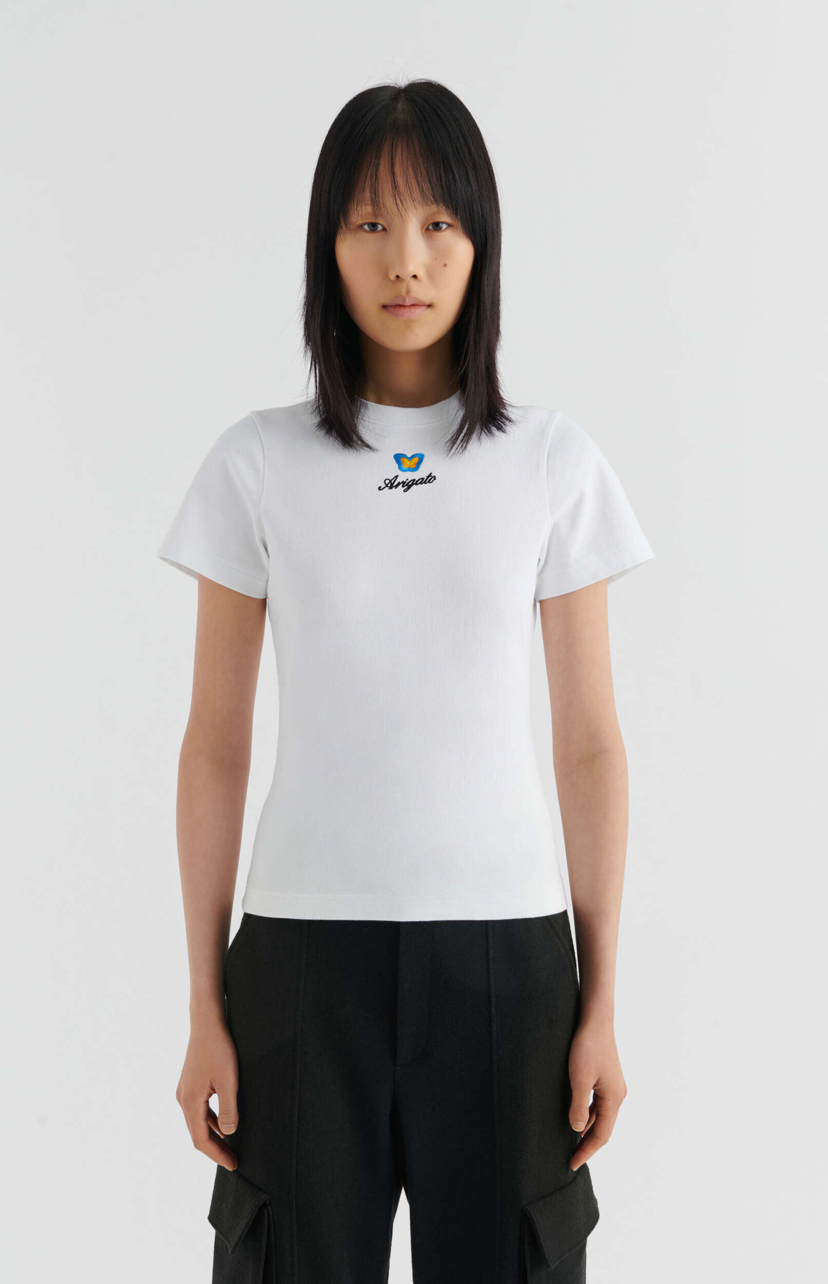 A full-length studio shot of a person with short black hair wearing a fitted white short-sleeve t-shirt that features a blue and yellow butterfly logo above the word 'Arigato' in black script, paired with black cargo pants. The person stands centered against a clean white background, looking directly at the camera.