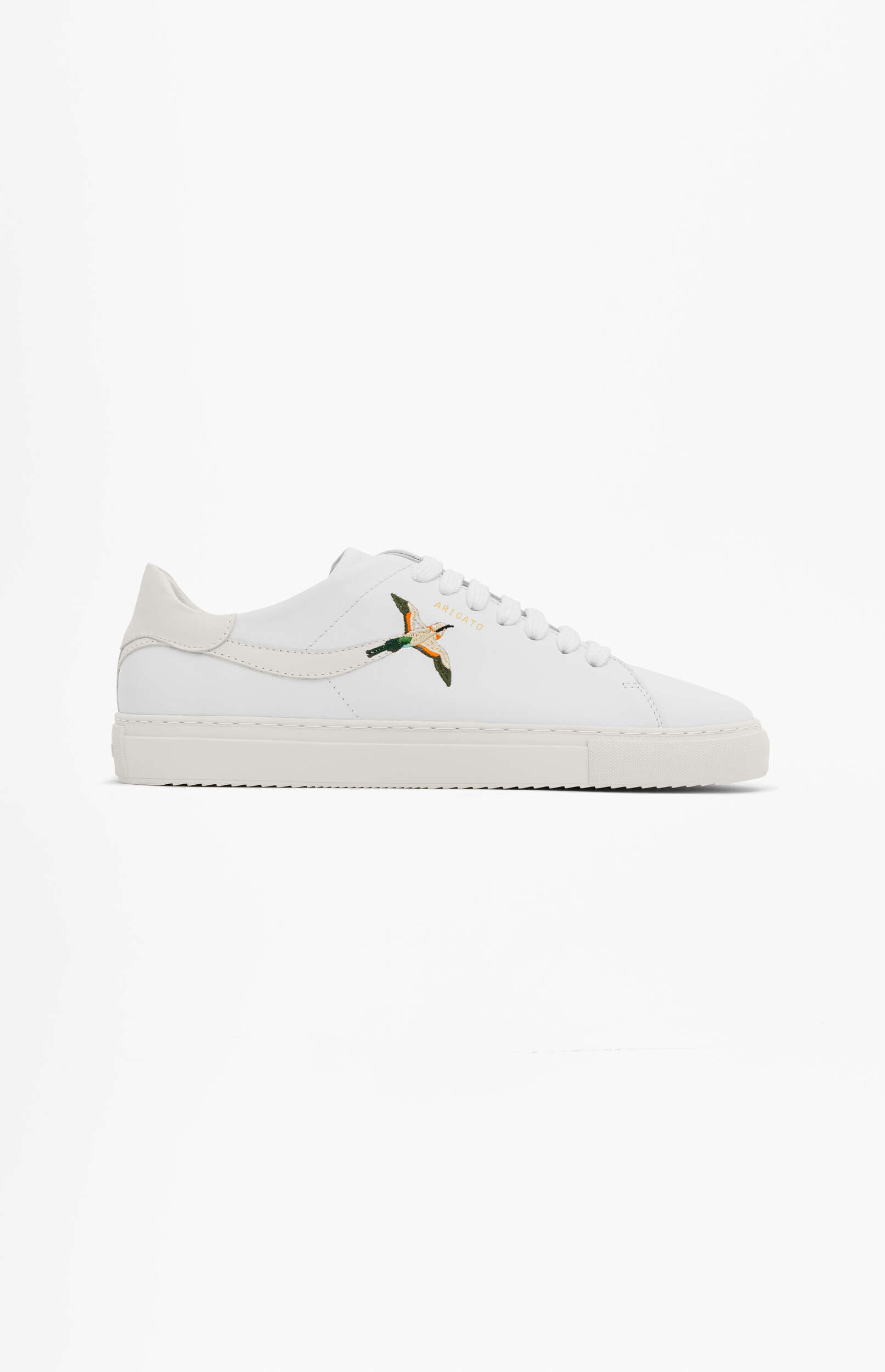 A white low-top sneaker with white laces and a white sole. An embroidered design of a flying bird, predominantly green and orange, is visible on the side, with the brand name 'ARIGATO' subtly placed above it. The shoe is presented on a plain white background.