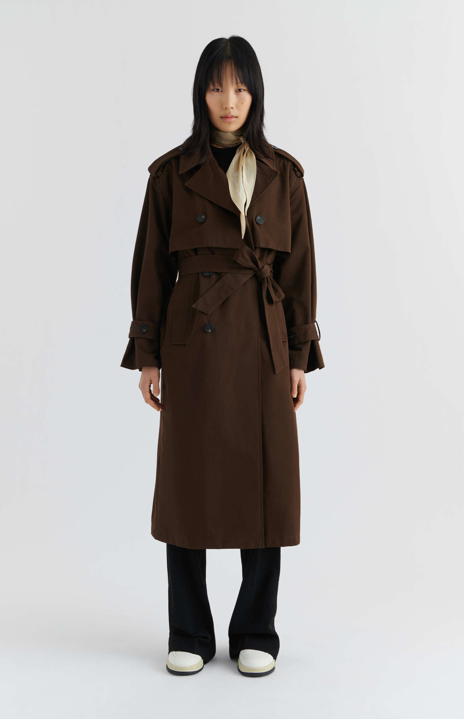 A full-length image of a person with dark, straight hair wearing a dark brown oversized trench coat, cinched at the waist, over black trousers, and white sneakers, accessorized with a light cream silk scarf, against a plain white background.