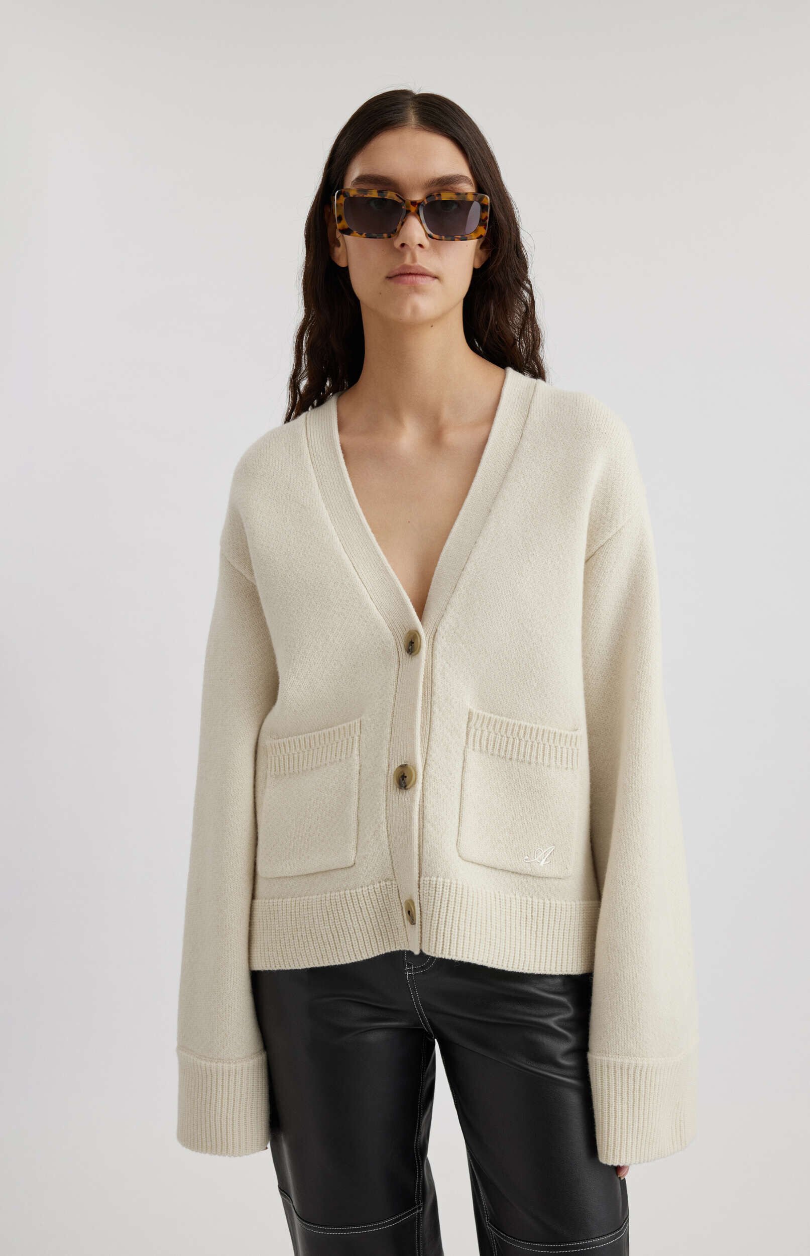 A woman wearing a cream-colored V-neck cardigan with two front pockets and brown buttons, black leather pants with white stitching, and tortoise-shell rectangular sunglasses stands against a white background.