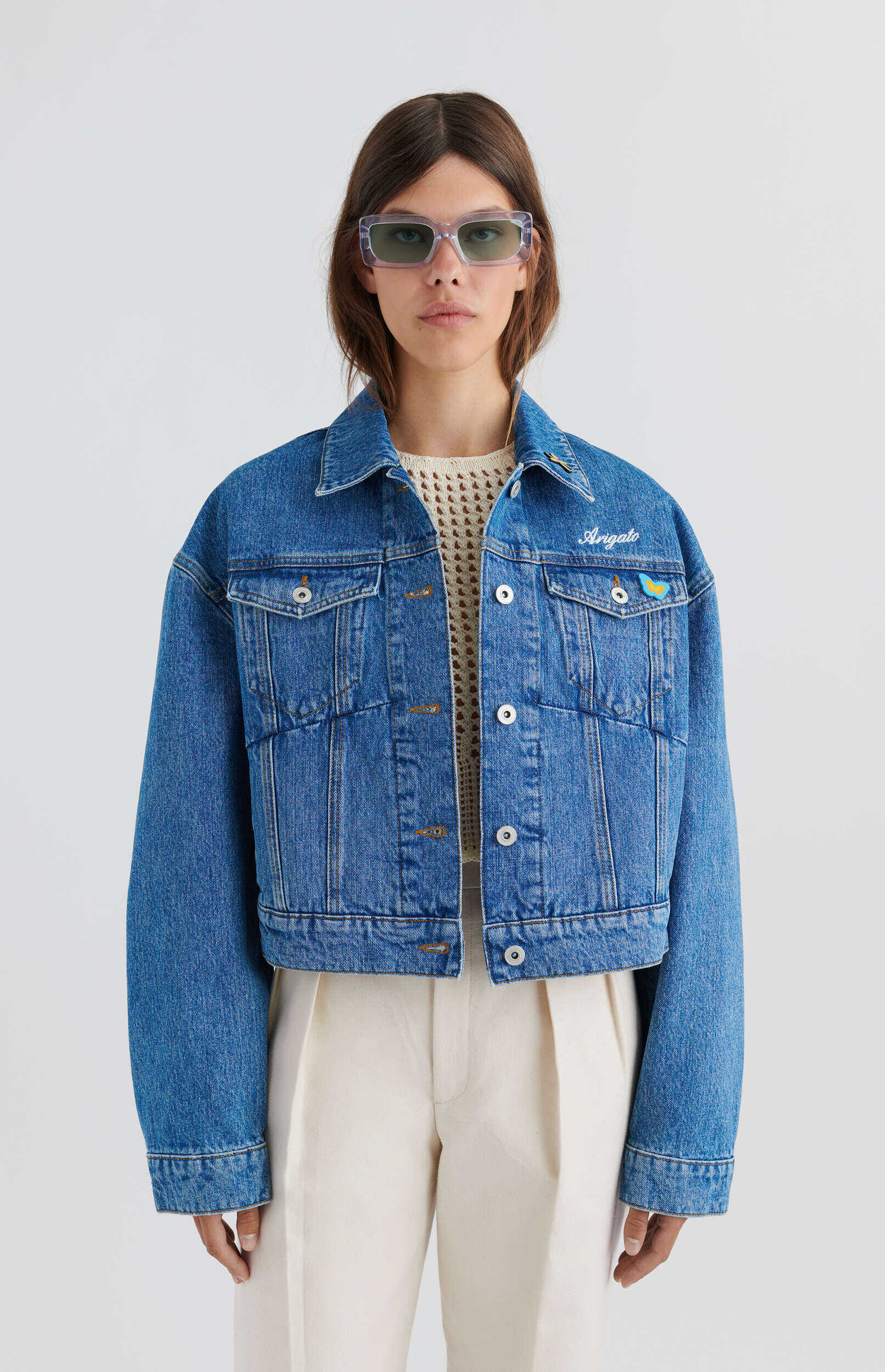 A woman stands against a white background, wearing a blue denim jacket with 'Arigato' embroidered in white script on the left chest and a small yellow and blue flag pin on the right chest pocket. She wears a cream-colored open-knit top underneath, cream trousers, and transparent lilac rectangular sunglasses.