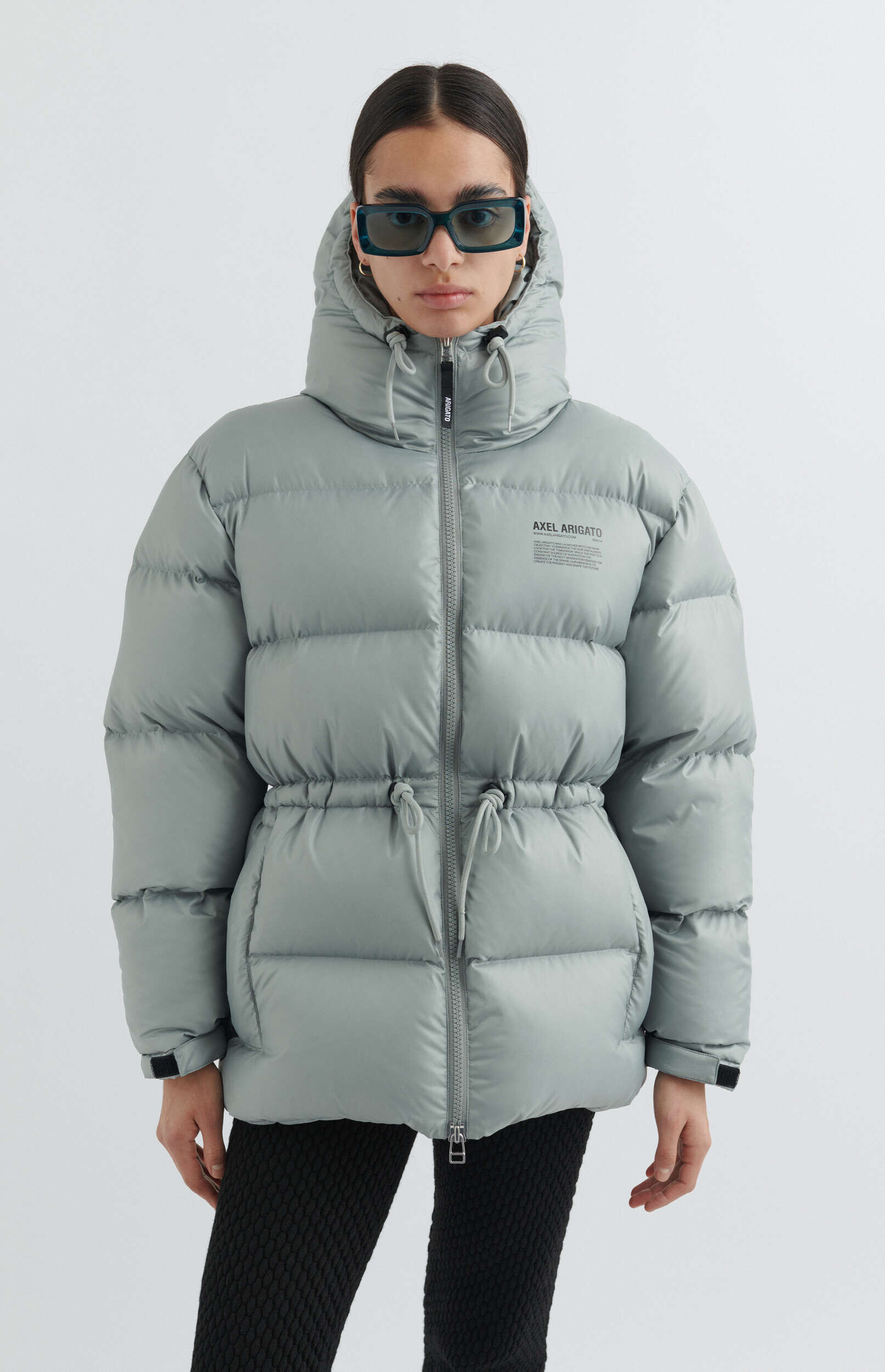 A person wearing a light grey hooded puffer jacket with a drawstring waist, dark rectangular sunglasses, and small gold hoop earrings, standing against a white background. The jacket features an 'AXEL ARIGATO' logo on the chest.