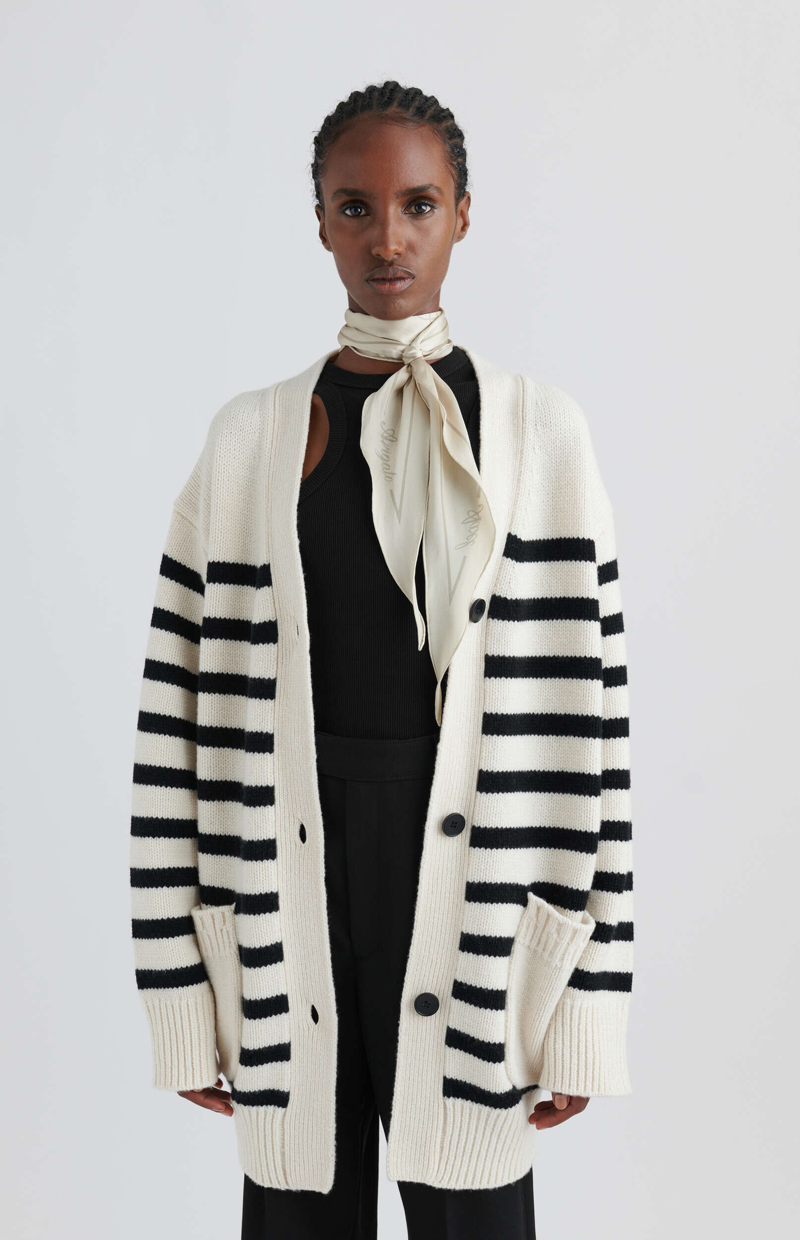 A Black woman with braided hair stands against a white background, wearing a stylish outfit. She is dressed in an oversized cream-colored cardigan with thick horizontal black stripes, layered over a black ribbed tank top with a shoulder cutout. A light beige silk scarf with subtle diagonal patterns is neatly tied around her neck. She also wears black pants, and the overall look is chic and sophisticated.
