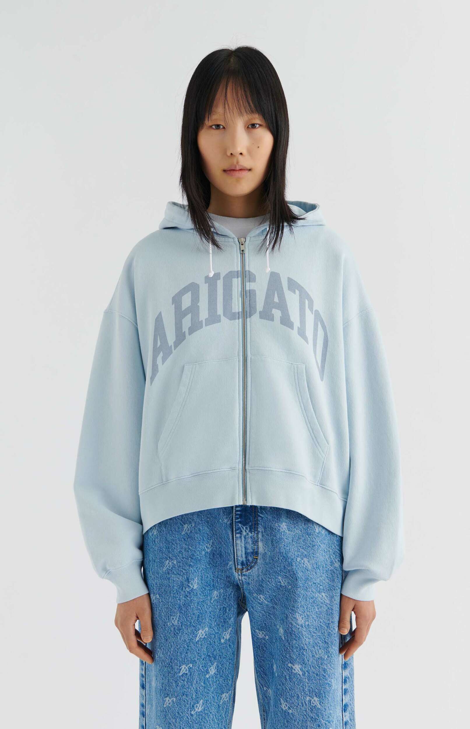A person with dark, straight hair and bangs, wearing a light blue zip-up hoodie with "ARIGATO" printed in an arched, darker blue-grey font on the chest, paired with light blue denim jeans featuring a subtle pattern, standing against a plain white background.
