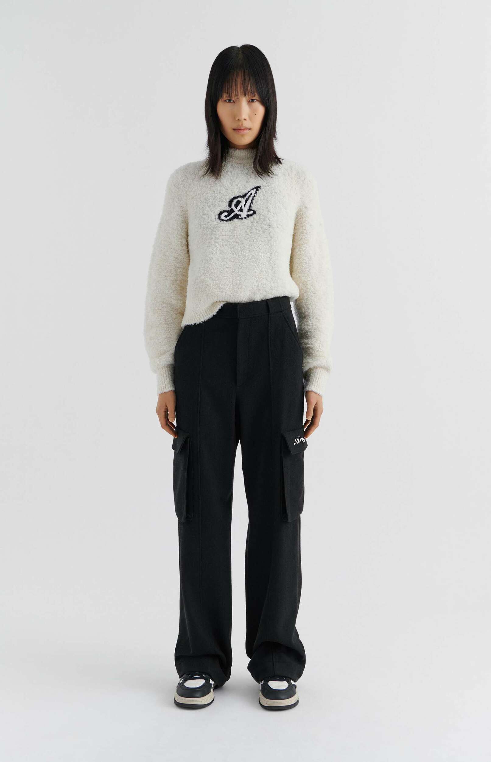 A person with dark, medium-length hair and bangs stands against a plain white background, wearing a cream-colored fuzzy sweater with a black cursive 'A' logo on the chest, black wide-leg cargo pants with pockets on the sides, and black and white sneakers.