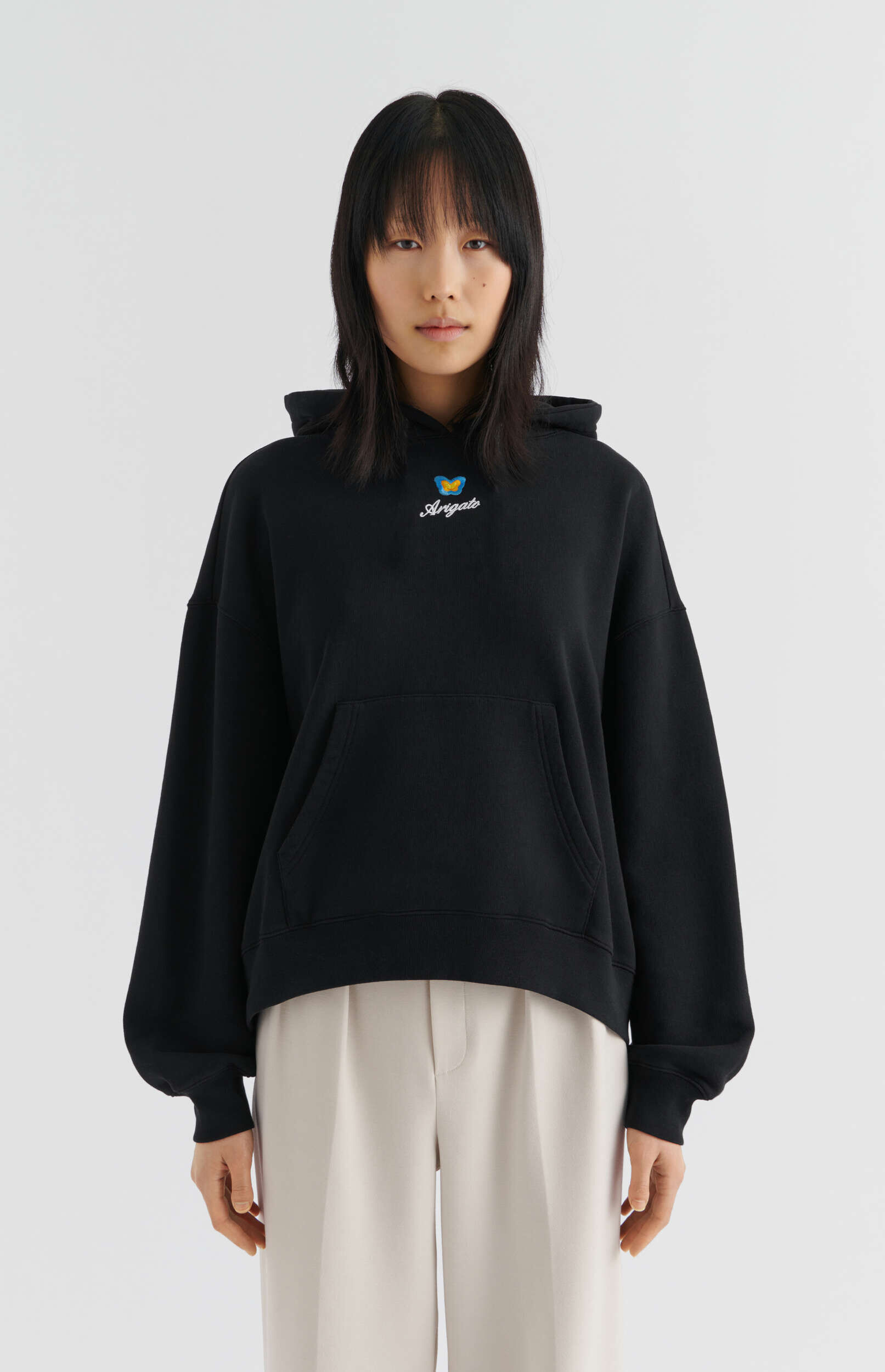 A person with dark hair and bangs wearing a black oversized hoodie featuring a blue and yellow butterfly logo and 'Arigato' text, paired with light beige wide-leg pleated trousers, standing against a white background.