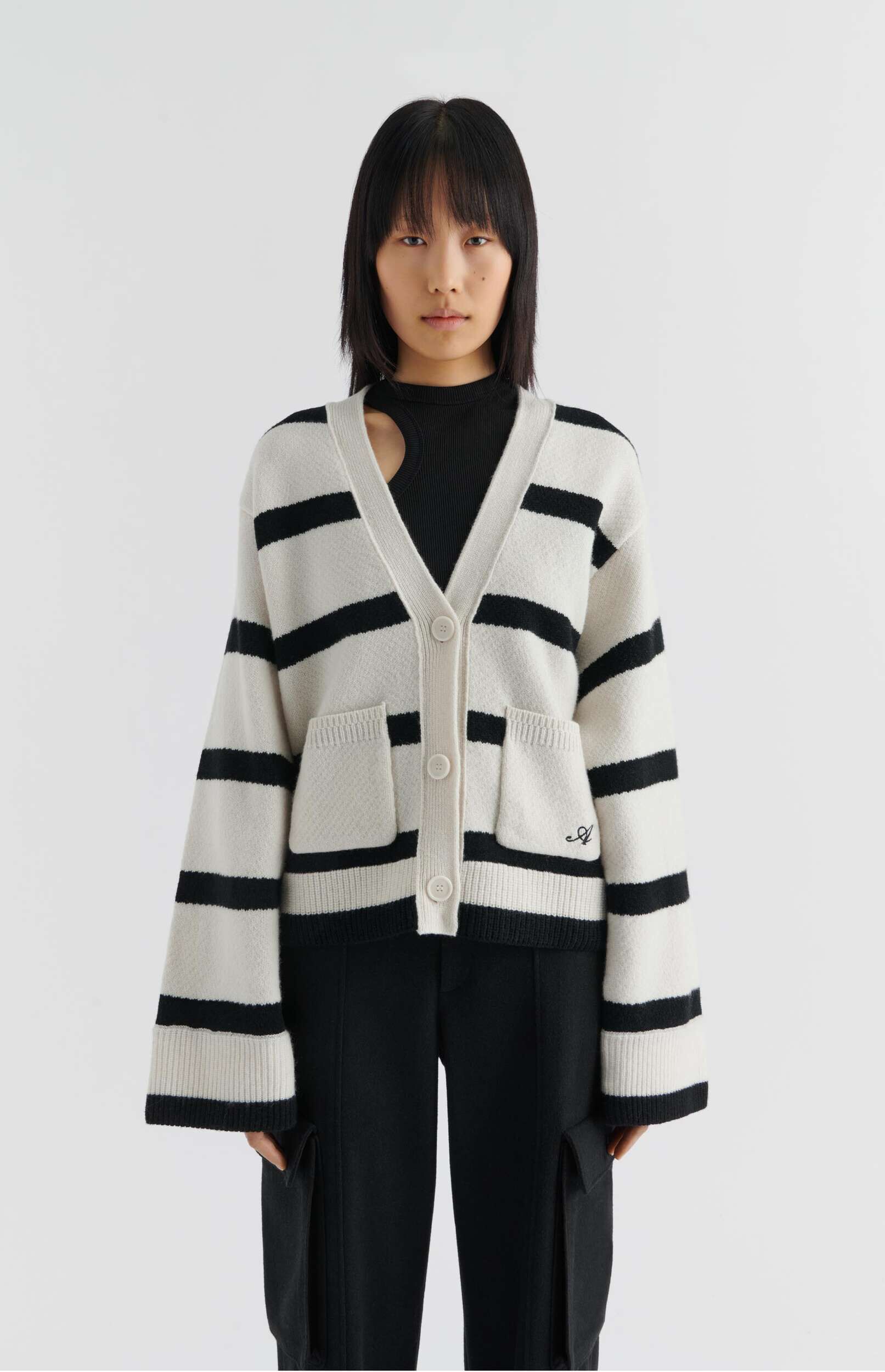 A young woman with dark hair and bangs wears a black and white striped V-neck cardigan over a black top with a shoulder cutout, paired with black cargo pants, standing against a white background.