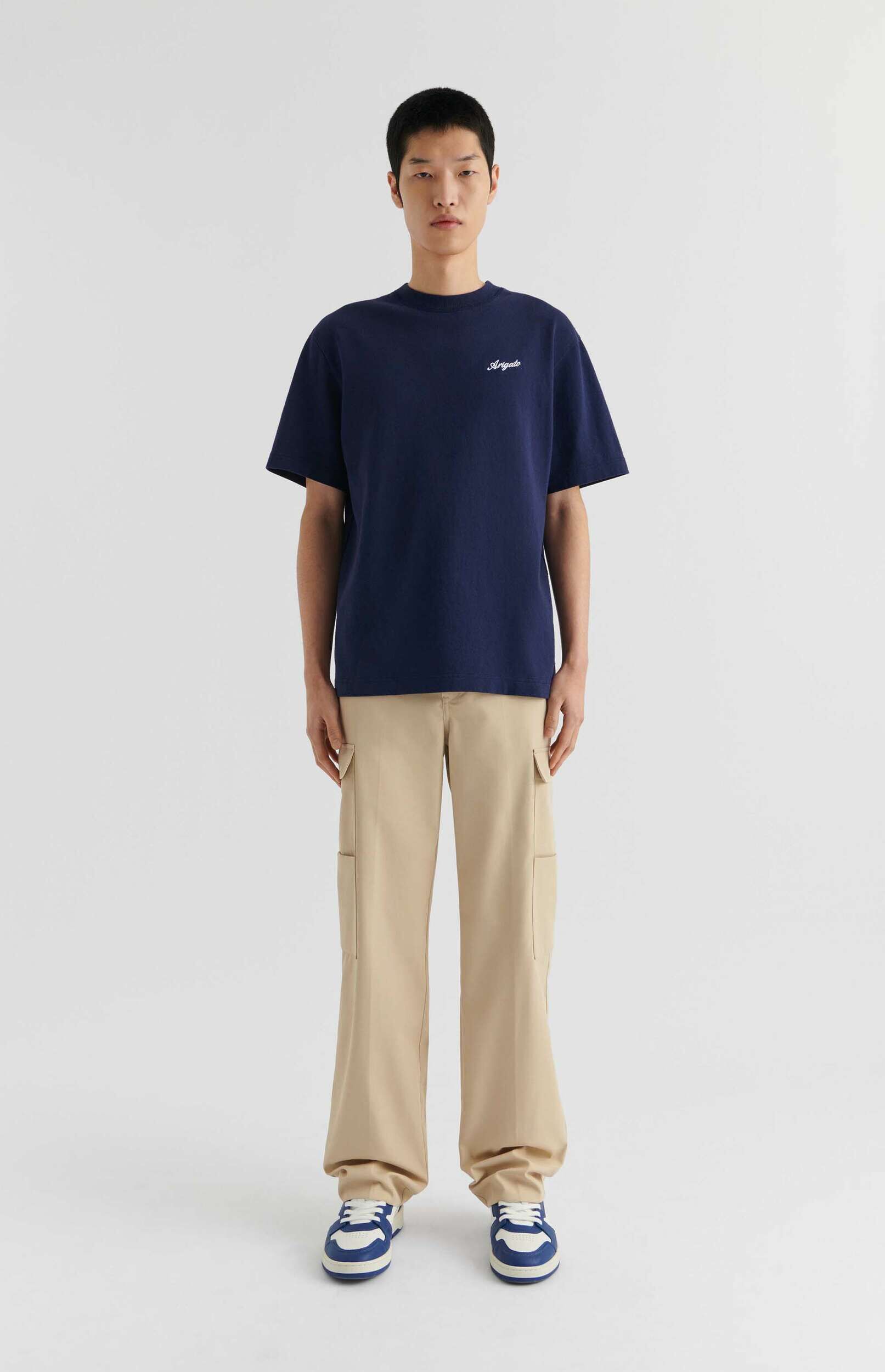 A full-body shot of a young man with short dark hair, standing against a plain white background. He is wearing a dark blue crew-neck t-shirt with 'Arigato' written in white script on the left chest, light beige relaxed-fit cargo pants, and blue and white sneakers. He stands still, looking directly forward.
