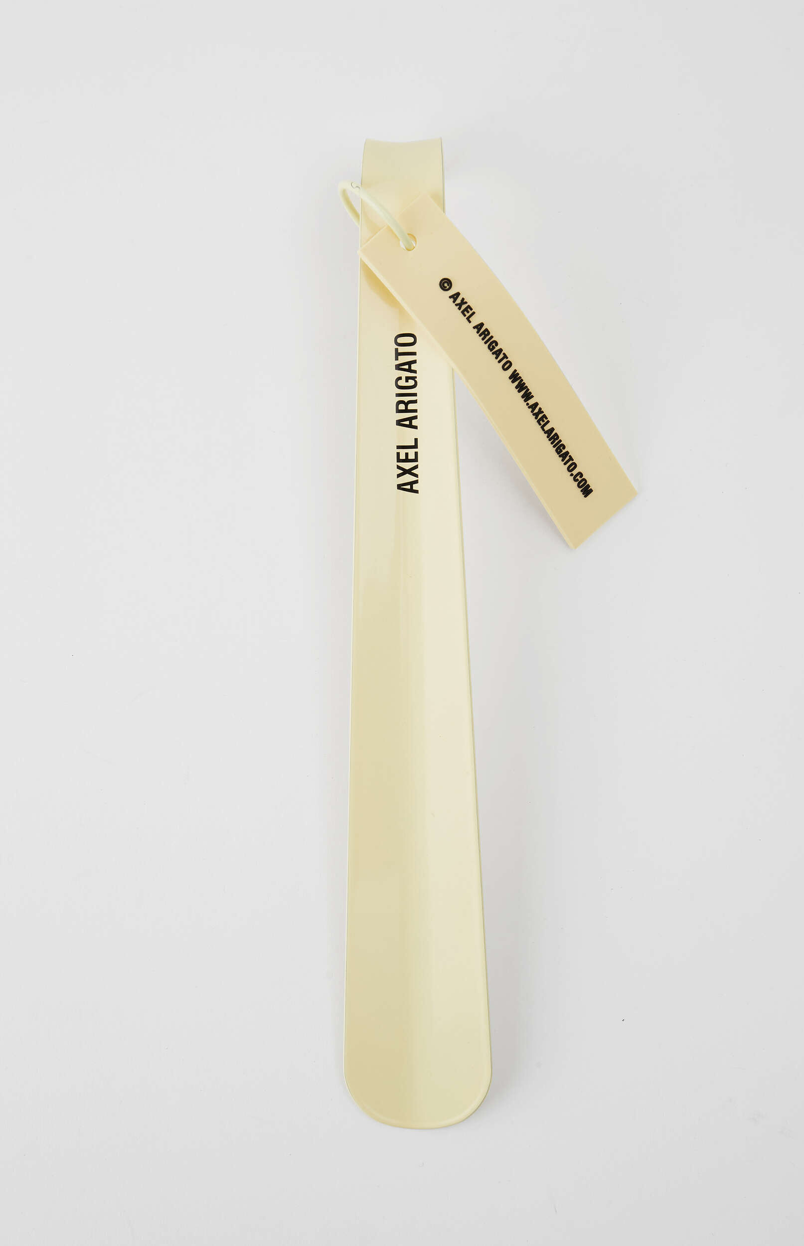 A light yellow or cream-colored Axel Arigato shoehorn stands upright on a white background, with a matching tag displaying the brand name and website attached to its top.