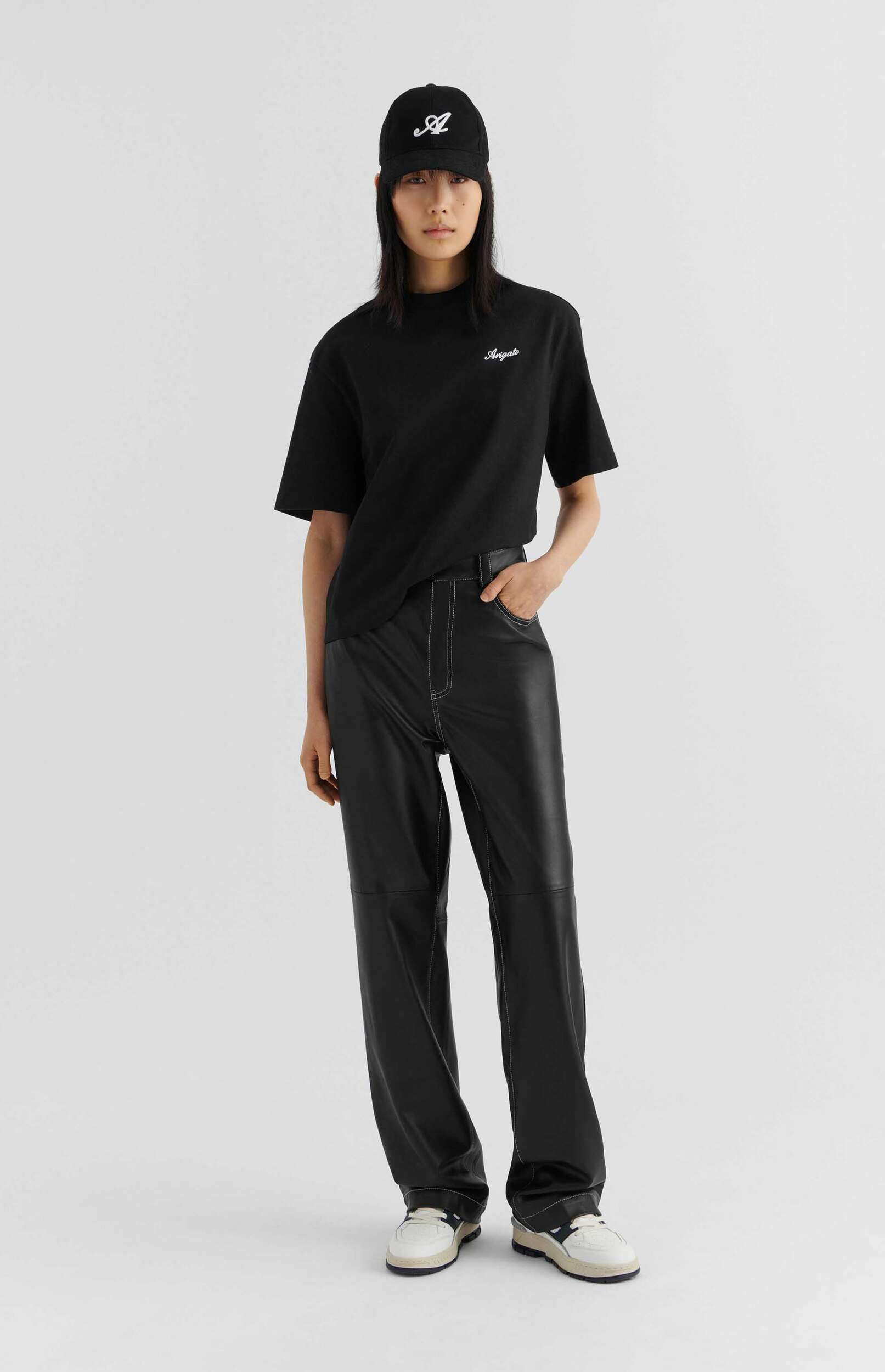 A person wearing a black t-shirt with 'Arigato' text, black leather pants with white contrast stitching, a black baseball cap with a white 'A' logo, and white and black sneakers, standing against a plain white background.